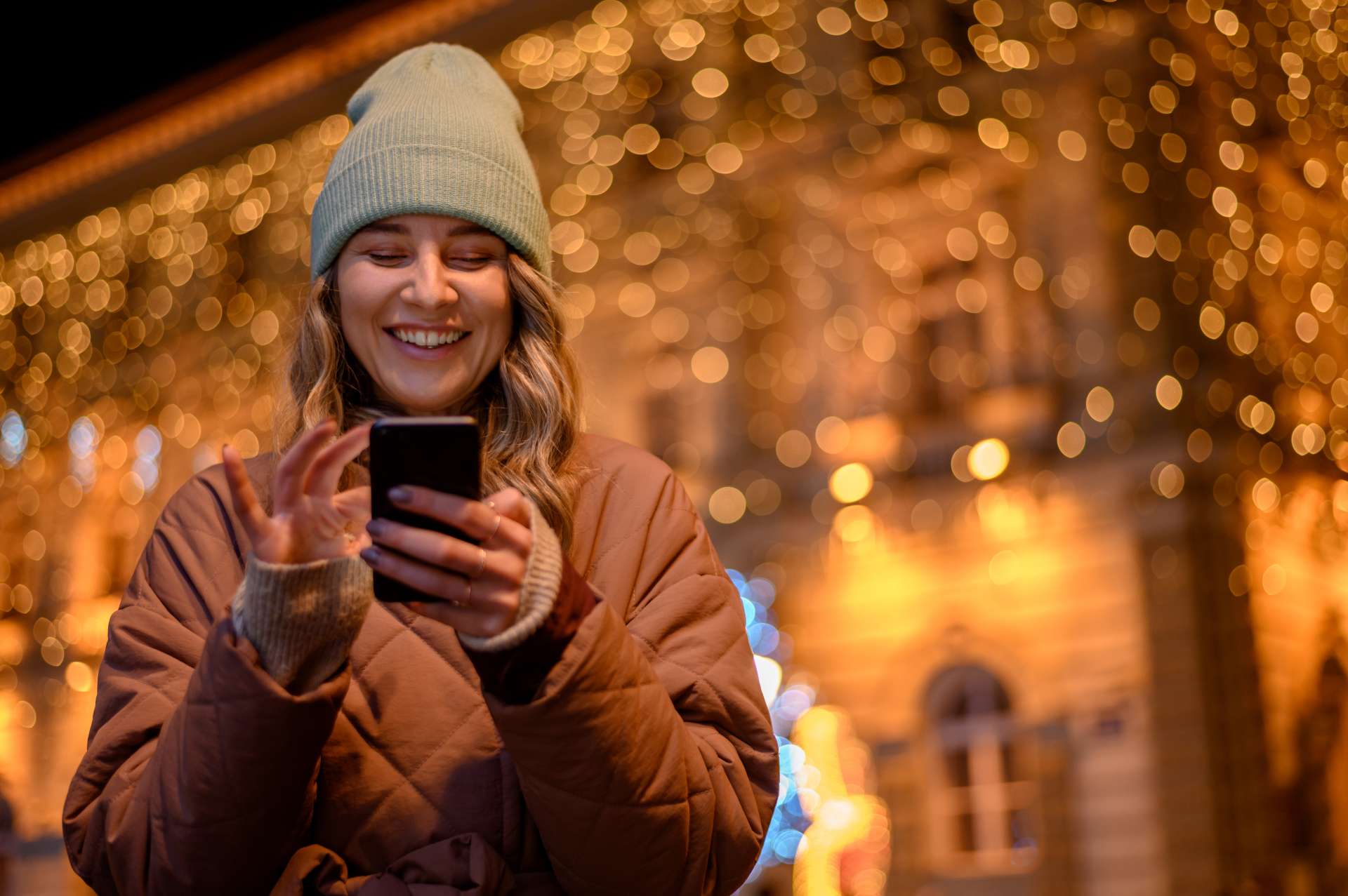 Woman in bobble hat outside a building with festive lights, smiling at her phone